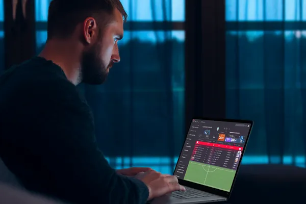 একজন পুরুষ তার ল্যাপটপে মনোযোগ দিয়ে কাজ করছেন, ব্যাকগ্রাউন্ডে লাইভ স্ট্যাটস — যা GOOD112 মোবাইল অ্যাপের স্পোর্টস ও গেমিং ফিচারগুলোকে তুলে ধরে।